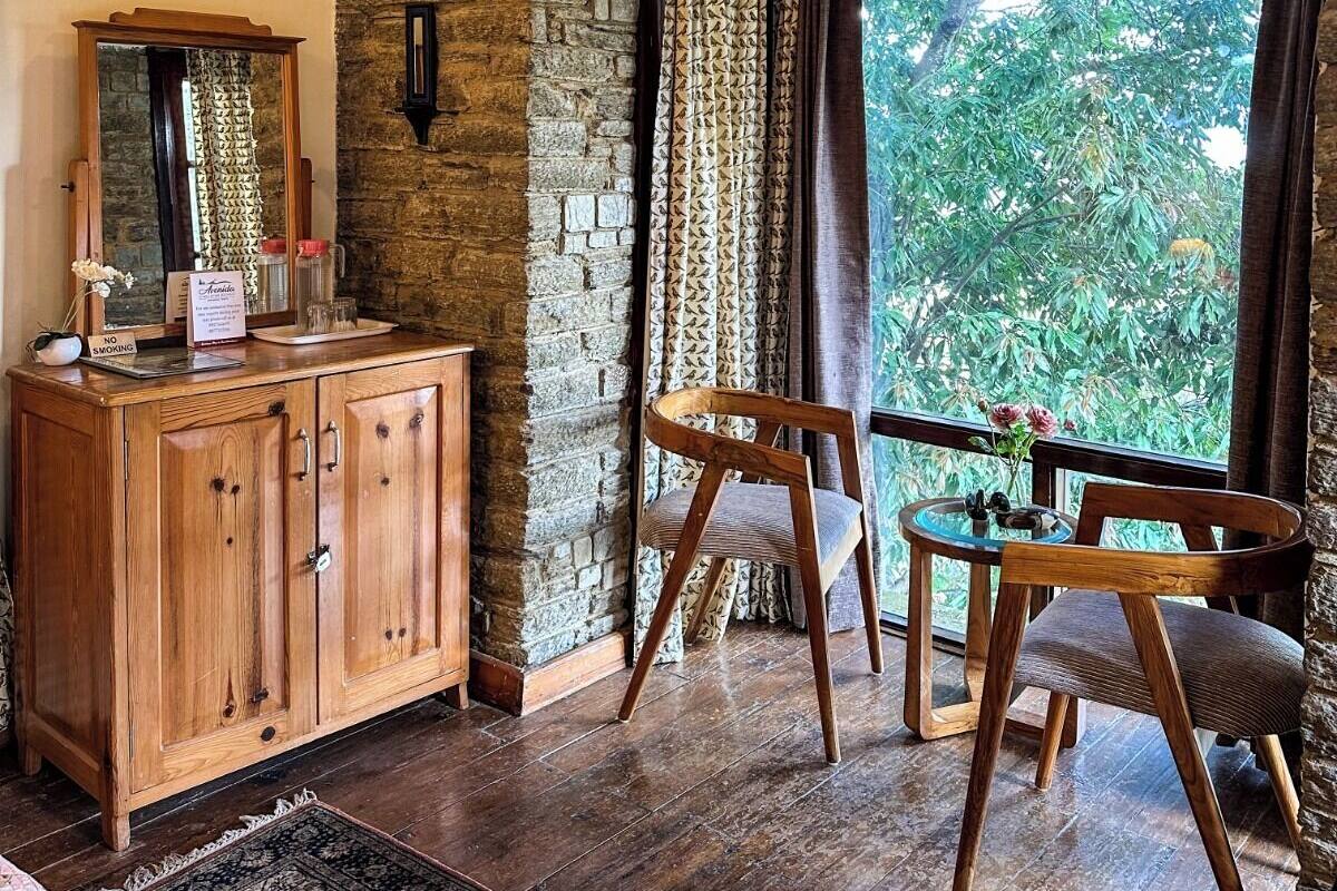 Une pièce rustique avec un mur en pierre, des poutres en bois au plafond, une armoire en bois, un miroir, une chaise, une petite table avec un vase, et une vue sur la verdure à l’extérieur.