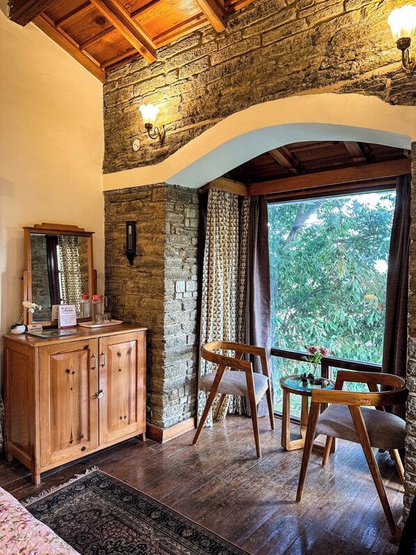 Una habitación rústica con pared de piedra, vigas de madera en el techo, un armario de madera, un espejo, una silla, una pequeña mesa con un jarrón y vistas a la vegetación.