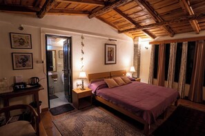 Un dormitorio acogedor con techo de madera, una cama con manta púrpura, dos lámparas de noche, una mesita de noche, un escritorio con cafetera y un cuadro en la pared.