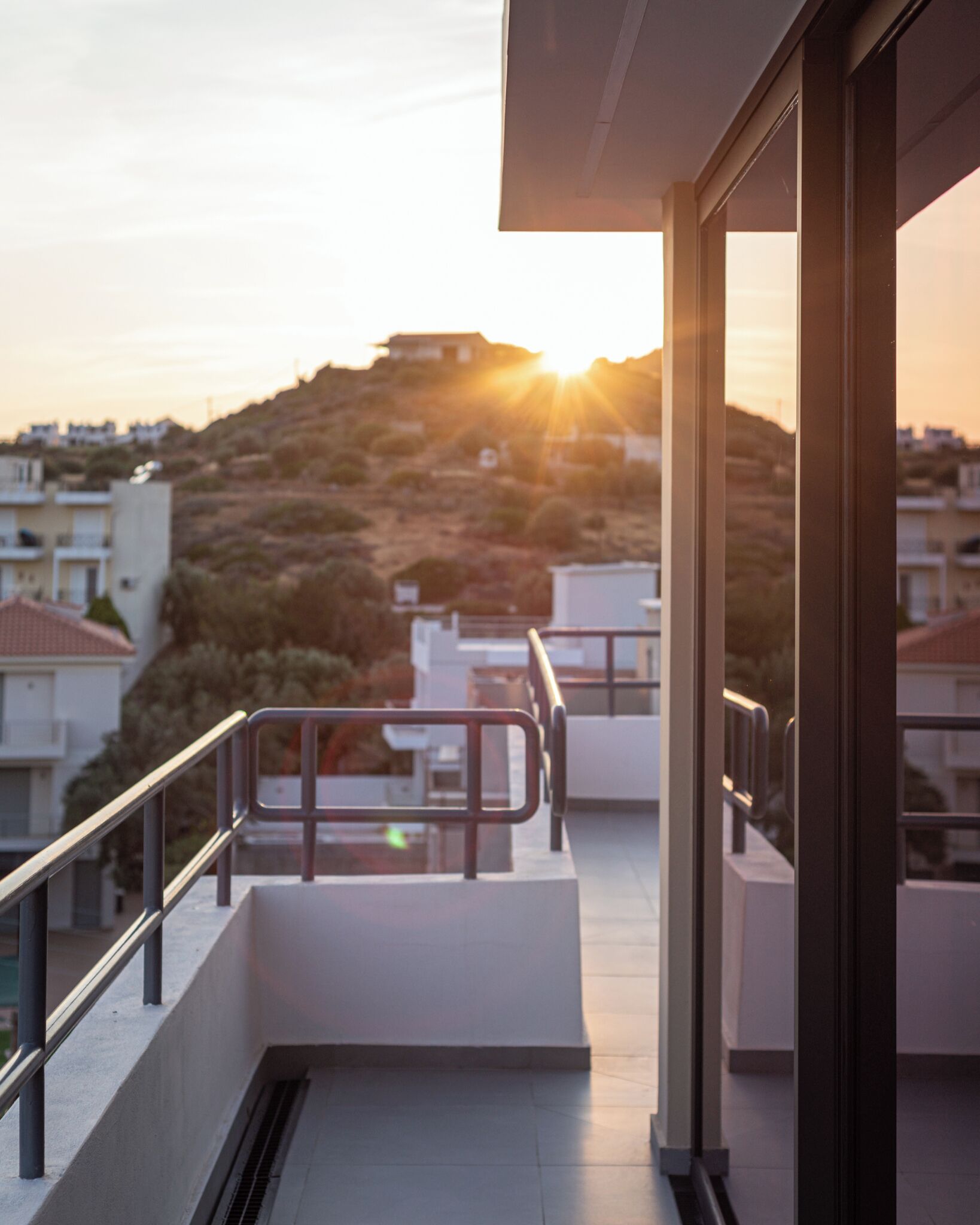 A balcony with a metal railing, a view of a hilly area with buildings, and the sun setting in the background.