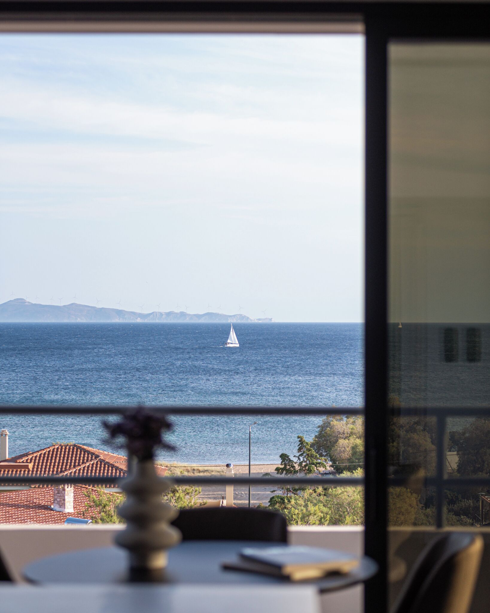 Ein Tisch mit einer Vase und einem Buch, ein Stuhl und Blick auf das Meer mit einem Segelboot und fernen Bergen.