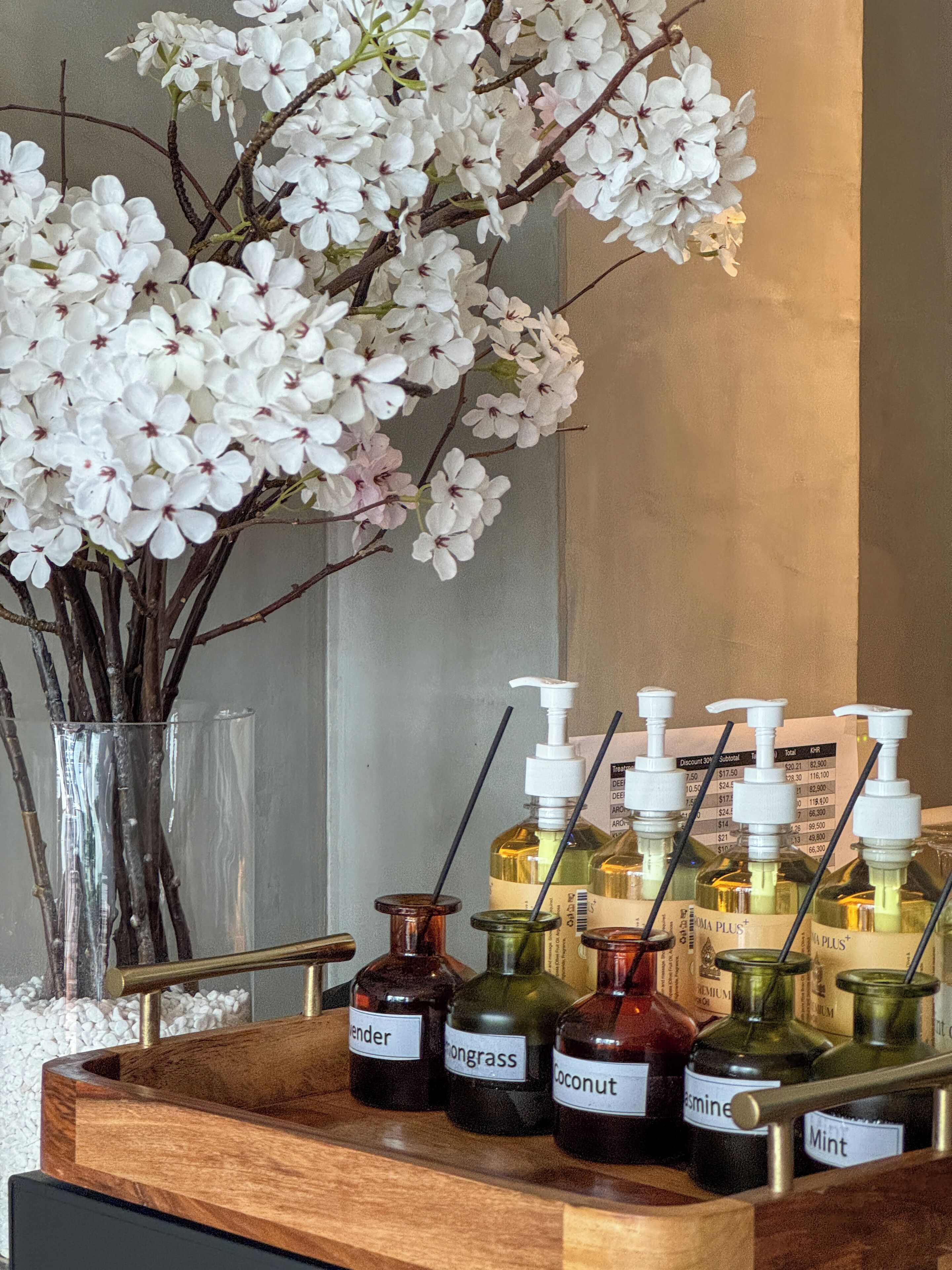 Un plateau en bois contenant différentes bouteilles d’huiles essentielles, chacune portant une étiquette, un vase avec des fleurs blanches et un miroir.