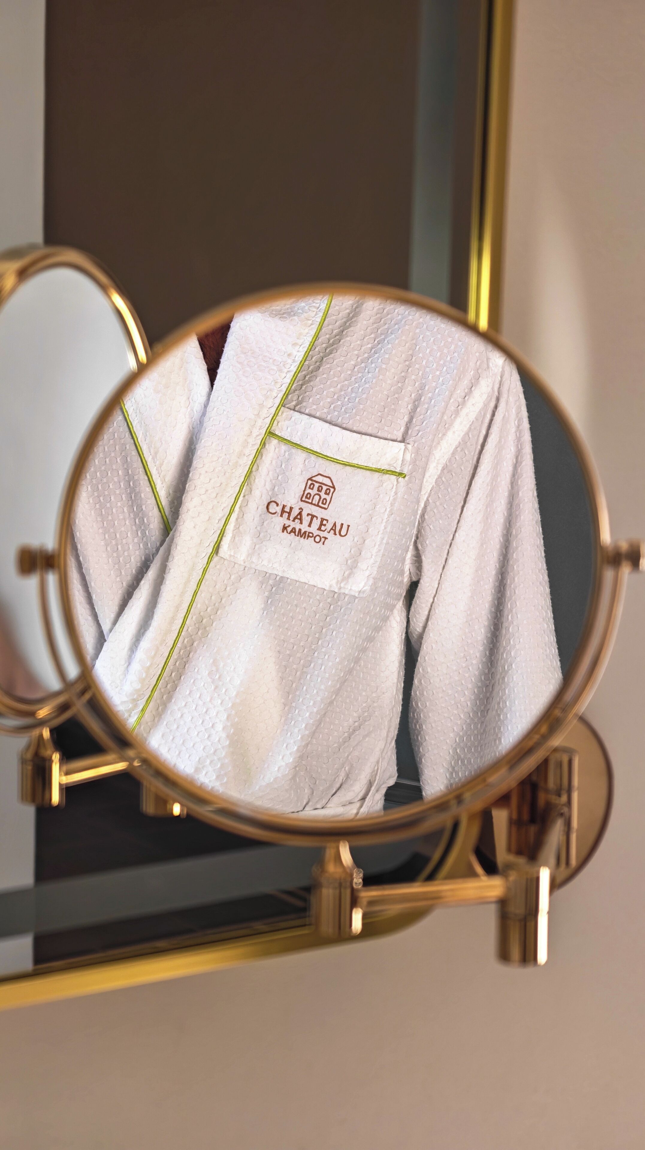 A round mirror with a gold frame reflecting a white robe with a label reading 'CHÂTEAU KAMPOT.'