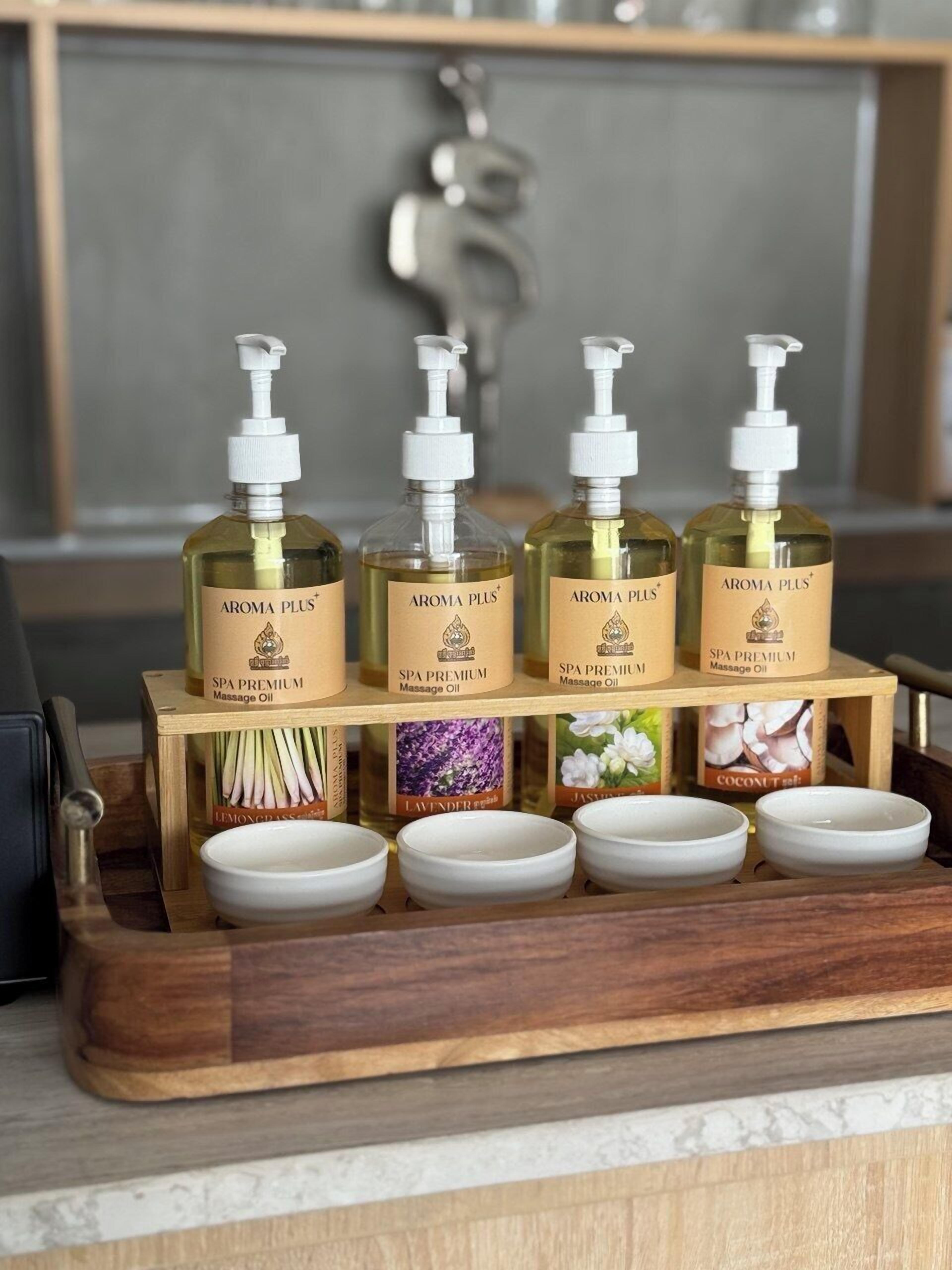 Un plateau en bois contenant quatre bouteilles d’huile de massage Aroma Plus et quatre petits bols remplis de substances de différentes couleurs.