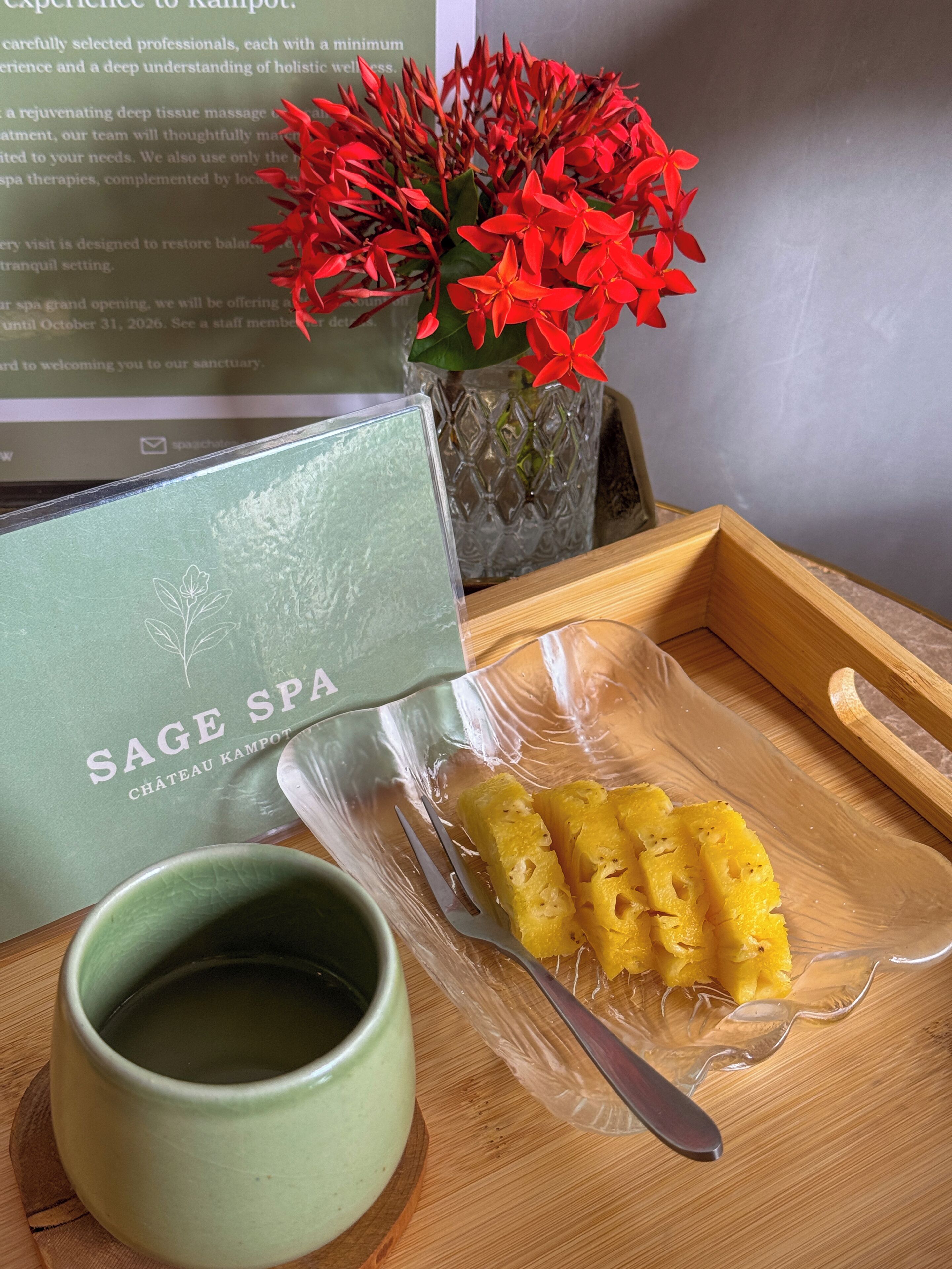 Una bandeja de madera con un recipiente de plástico que contiene rodajas de piña, una taza verde, un florero con flores rojas y un folleto de Sage Spa.