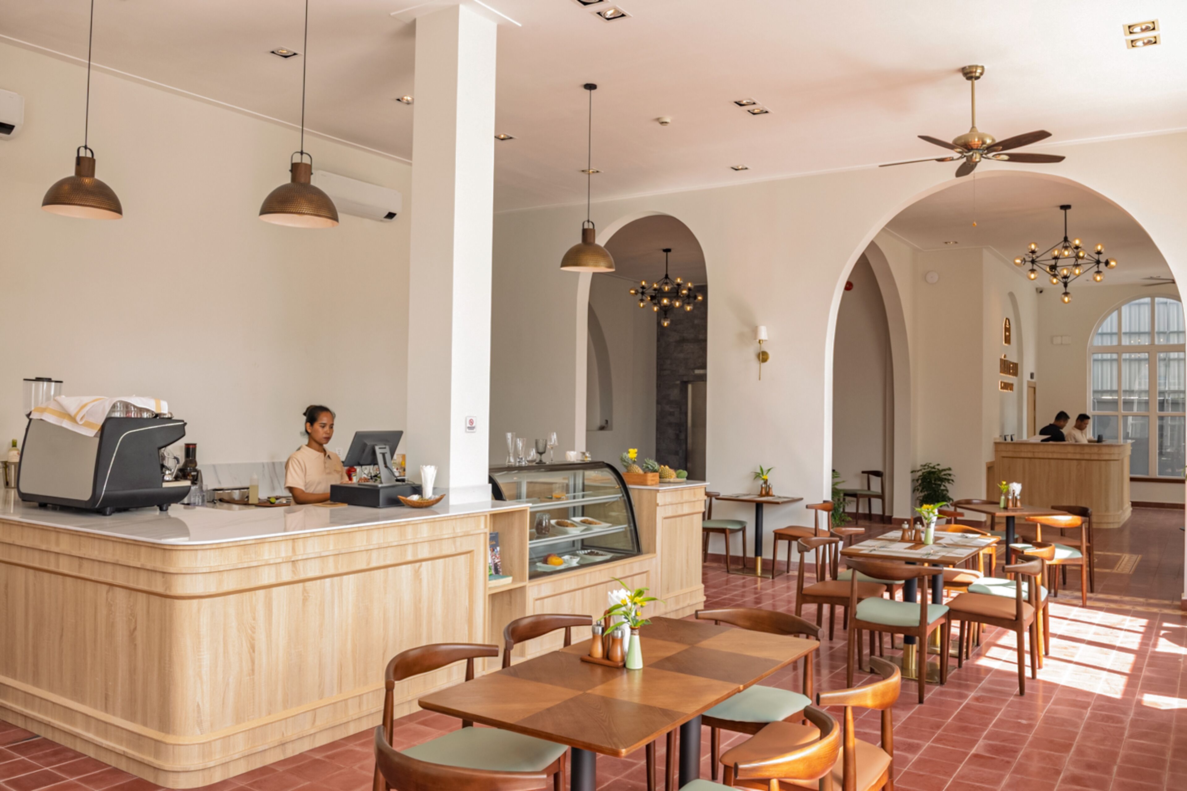A spacious cafe with wooden tables and chairs, a central counter with a coffee machine, and a ceiling fan.