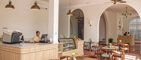 Una cafeteria àmplia amb taules i cadires de fusta, una barra central amb una màquina de cafè i un ventilador de sostre.