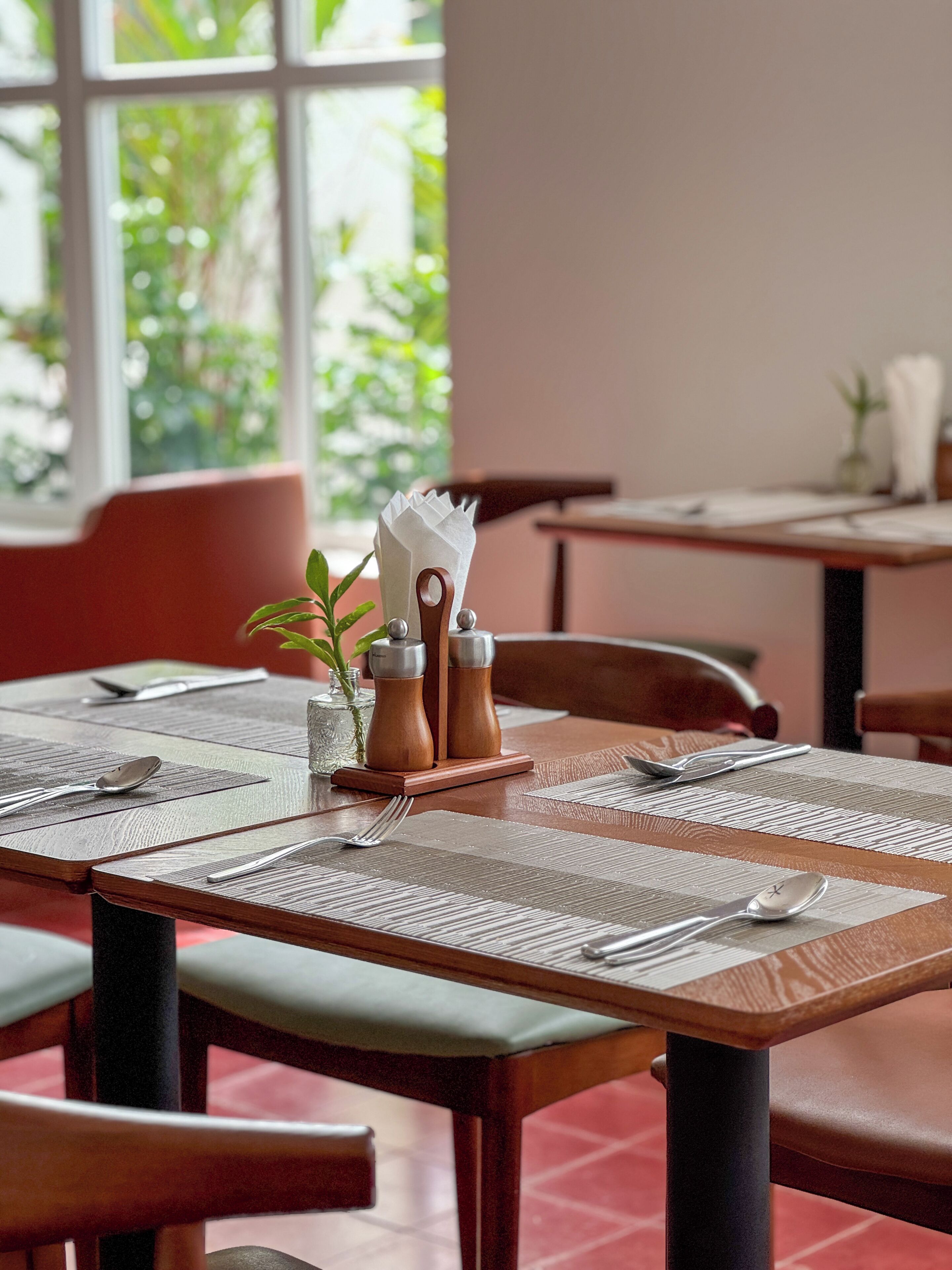 A dining area with tables set for two, featuring wooden chairs and a window with a view of greenery.