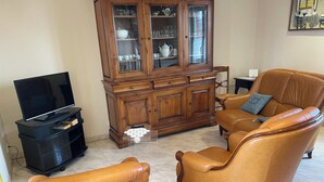 Een woonkamer met een televisie, leren fauteuils, een houten kast met glazen deuren en een betegelde vloer.