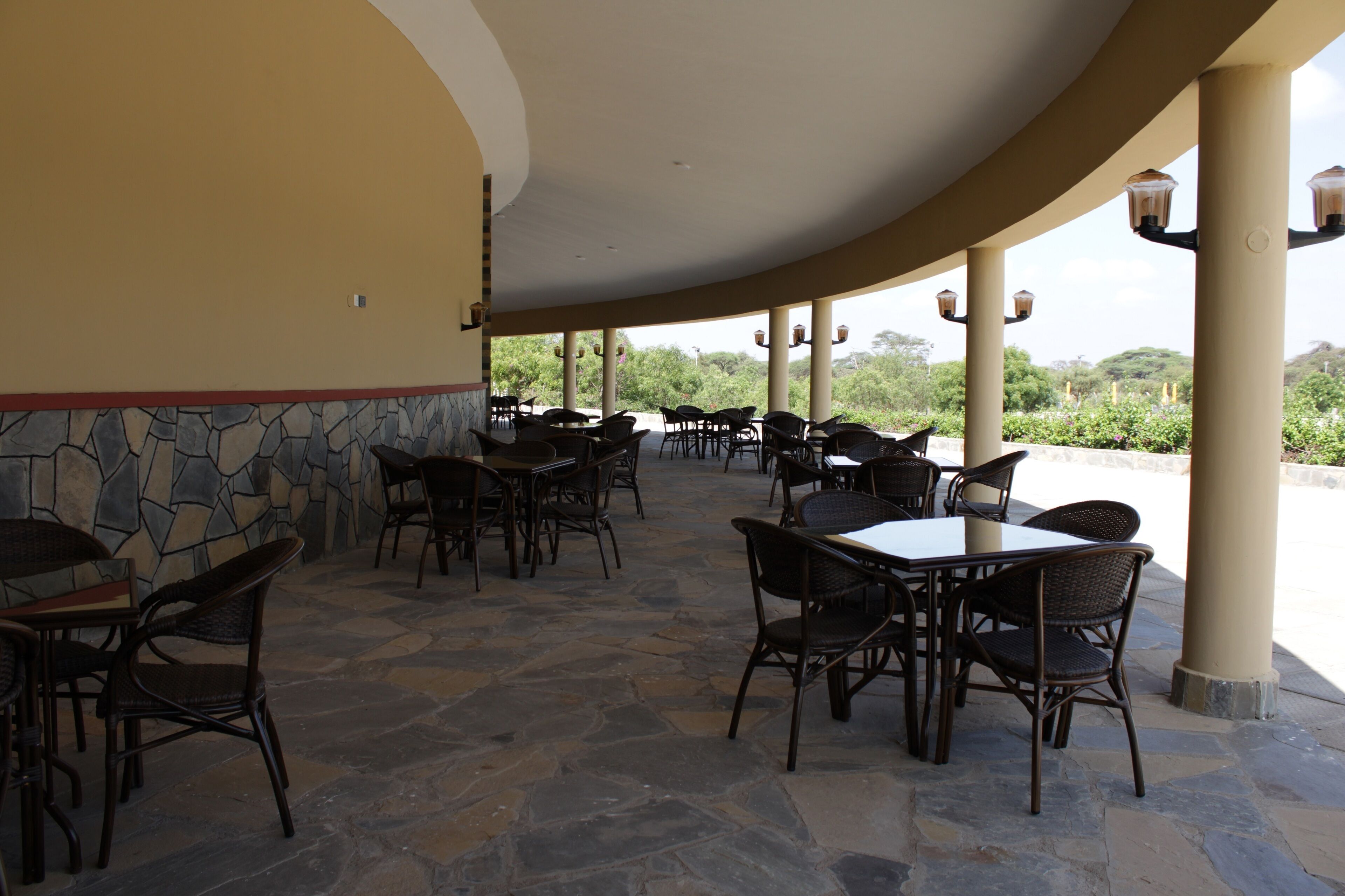 An outdoor patio with stone flooring, tables, and wicker chairs, surrounded by a low stone wall and columns with hanging lights.