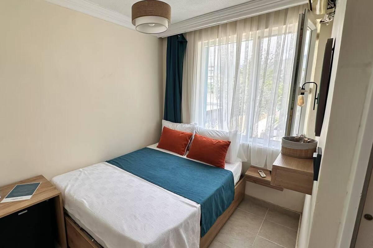 A hotel room with a bed, a bedside table with a lamp, a window with sheer curtains, and a view of the outdoors.