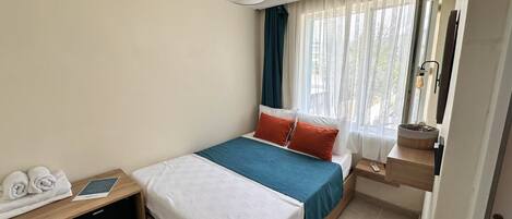 Ein Hotelzimmer mit einem Bett, einem Nachttisch mit Lampe, einem Fenster mit leichtem Vorhang und Blick ins Freie.
