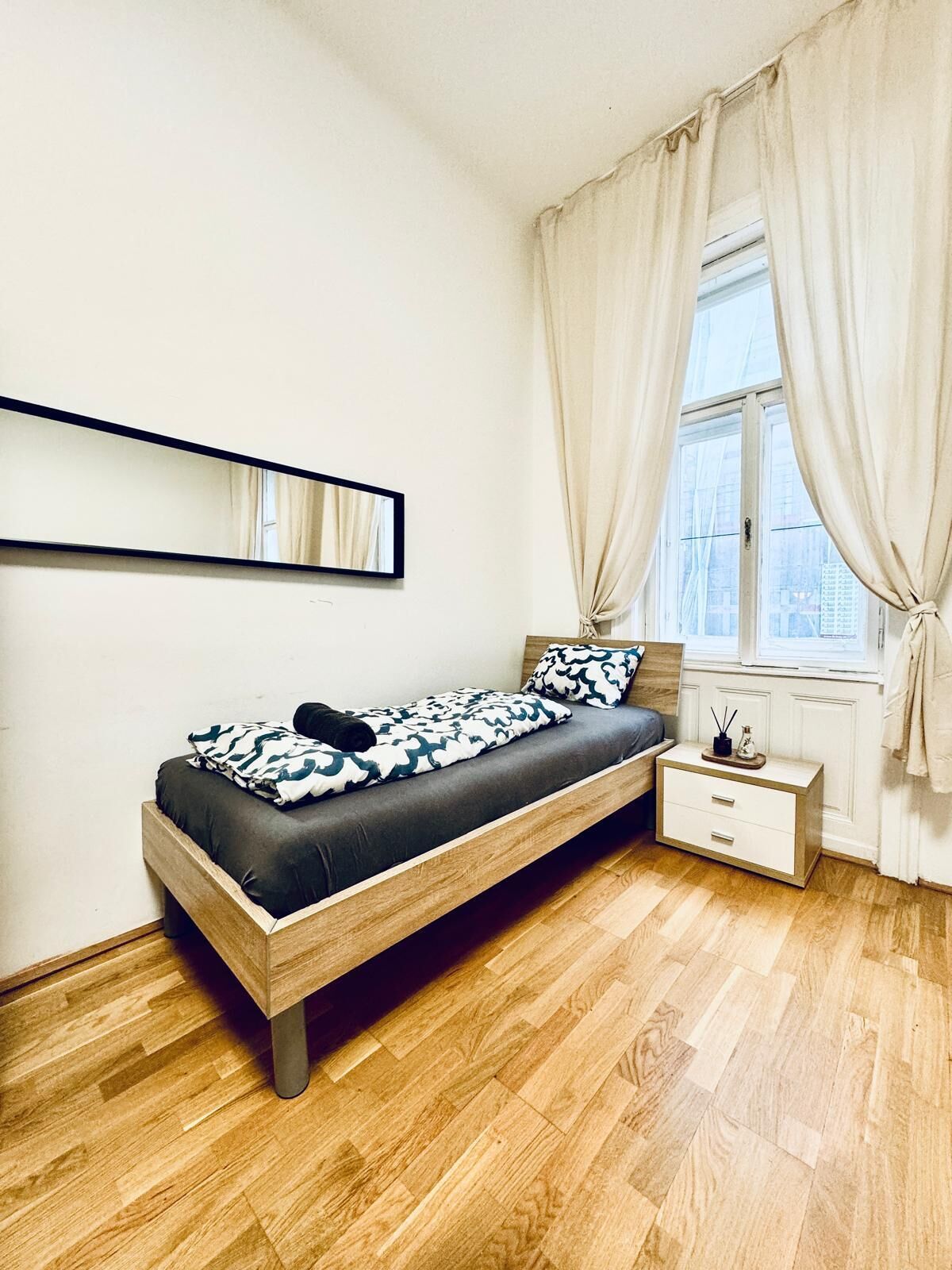 Een slaapkamer met een bed, nachtkastje, spiegel en een raam met gordijnen.
