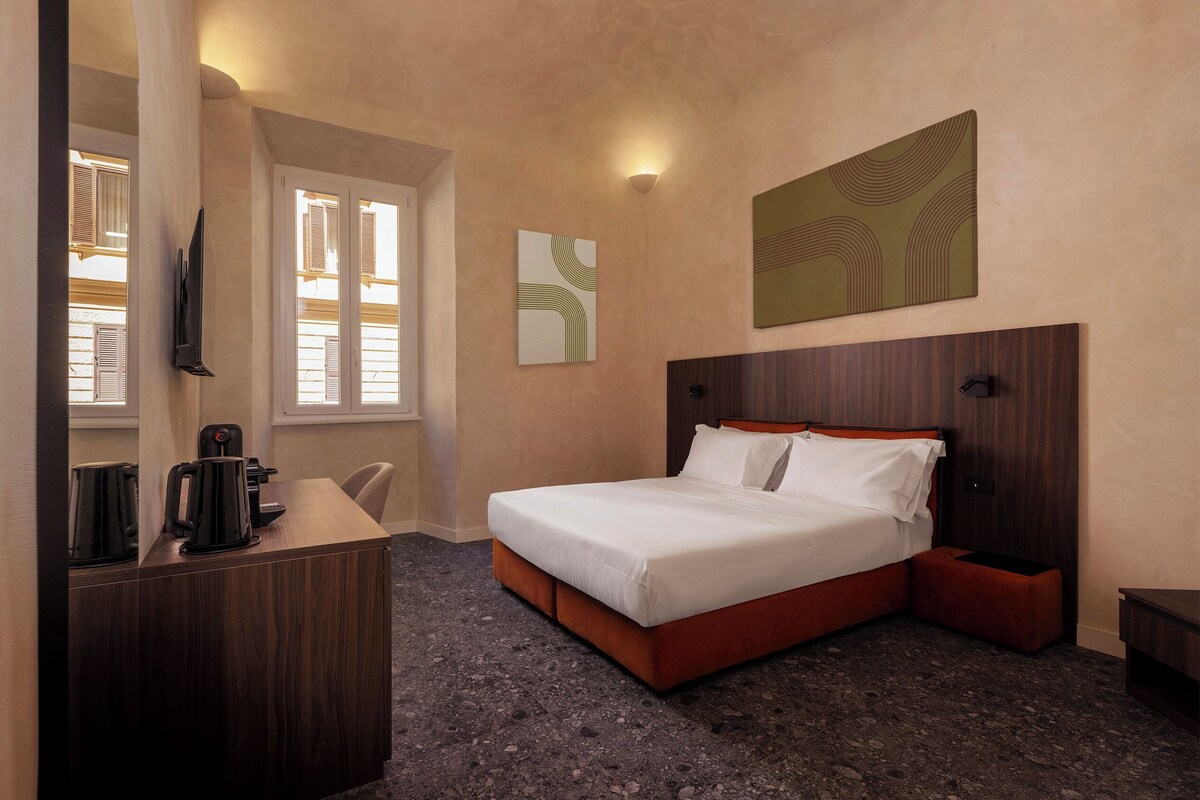 A hotel room with a bed, a desk with a coffee maker, a chair, and artwork on the walls.