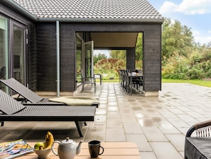 Ein überdachter Außenbereich mit Liegestühlen und einem Tisch, Blick auf einen Garten und eine Terrasse mit gefliestem Boden.
