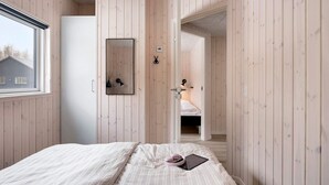 Ein Schlafzimmer mit Holzvertäfelung, einem Bett mit weißer Bettdecke, einem Nachttisch mit Lampe und einem Fenster mit Blick auf ein Haus.