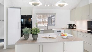 Eine moderne Küche mit weißen Schränken, einer großen Kochinsel mit Obstschale, zwei Blumentöpfen und einem Fenster, das für natürliches Licht sorgt.