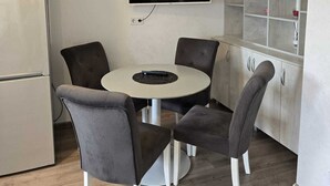 Un espacio habitable compacto con kitchenette, un comedor con una mesa redonda y cuatro sillas, un televisor de pantalla plana montado en la pared y un mueble empotrado con algunos objetos.