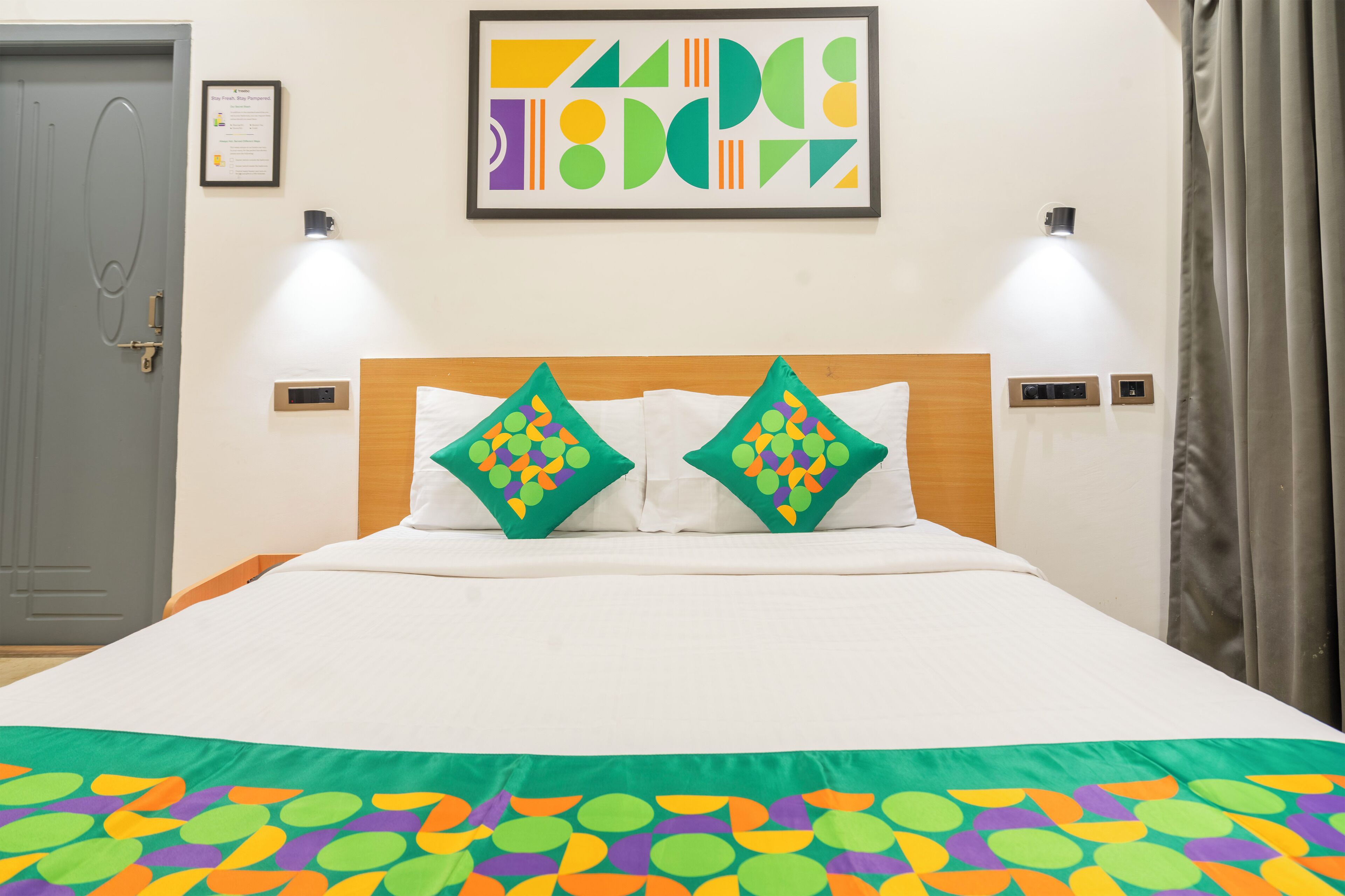 Une chambre d’hôtel comprenant un lit, deux oreillers à motifs géométriques, une œuvre d’art abstraite encadrée, deux lampes fixées au mur et une porte avec une poignée.