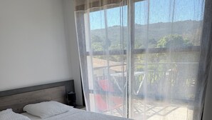 Un dormitorio con una cama grande, cabecera, mesita de noche y vistas al exterior a través de cortinas traslúcidas.