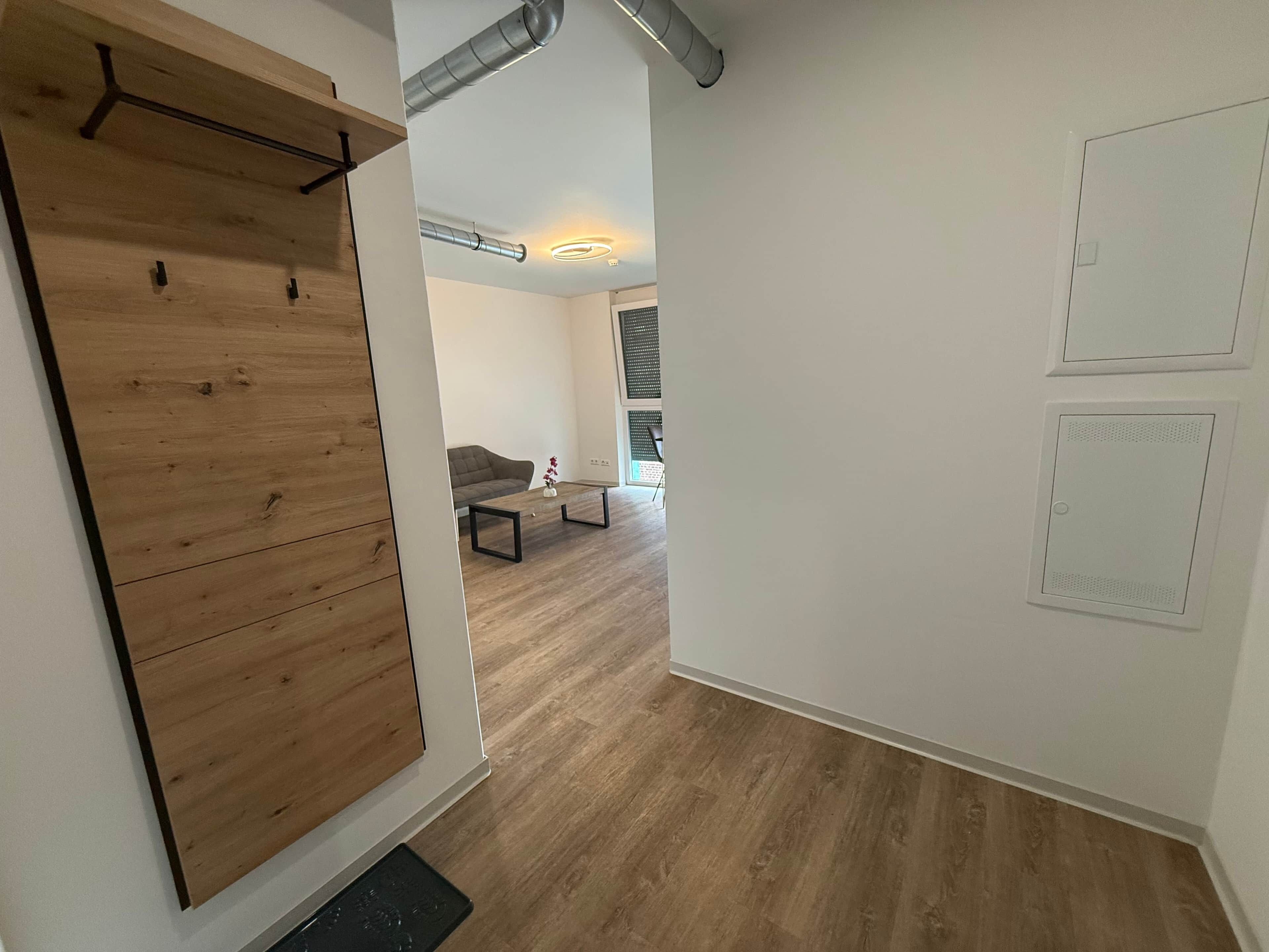 A modern living room with a wooden floor, a grey sofa, and a coffee table. There is a wooden wardrobe with a black handle, a white wall with two ventilation grilles, and a ceiling with exposed pipes.