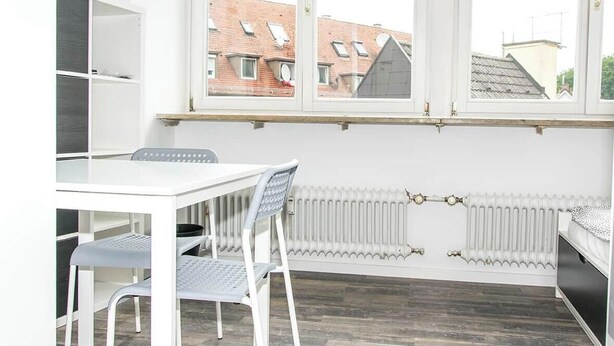 A room with a white table and chairs, a radiator, and a window with a view of buildings. - 15 - Bachwiesenstr. Stuttgart (Stuttgart)