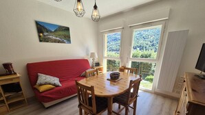 A cozy room with a red sofa, wooden dining table, and chairs, large windows with a view of greenery, and a painting on the wall. - Cozy apartment in Cauterets with parking and wifi (Cauterets)