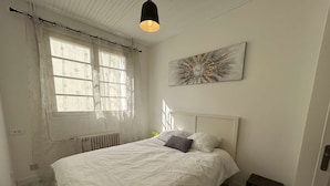 Une chambre à coucher comprenant un lit, un radiateur, une fenêtre avec des rideaux, une œuvre d’art accrochée au mur et une porte.