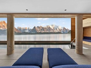 Ein Spa-Raum mit zwei blauen Liegestühlen, einer Holzverkleidung und einem großen Fenster mit Blick auf Berge und See.