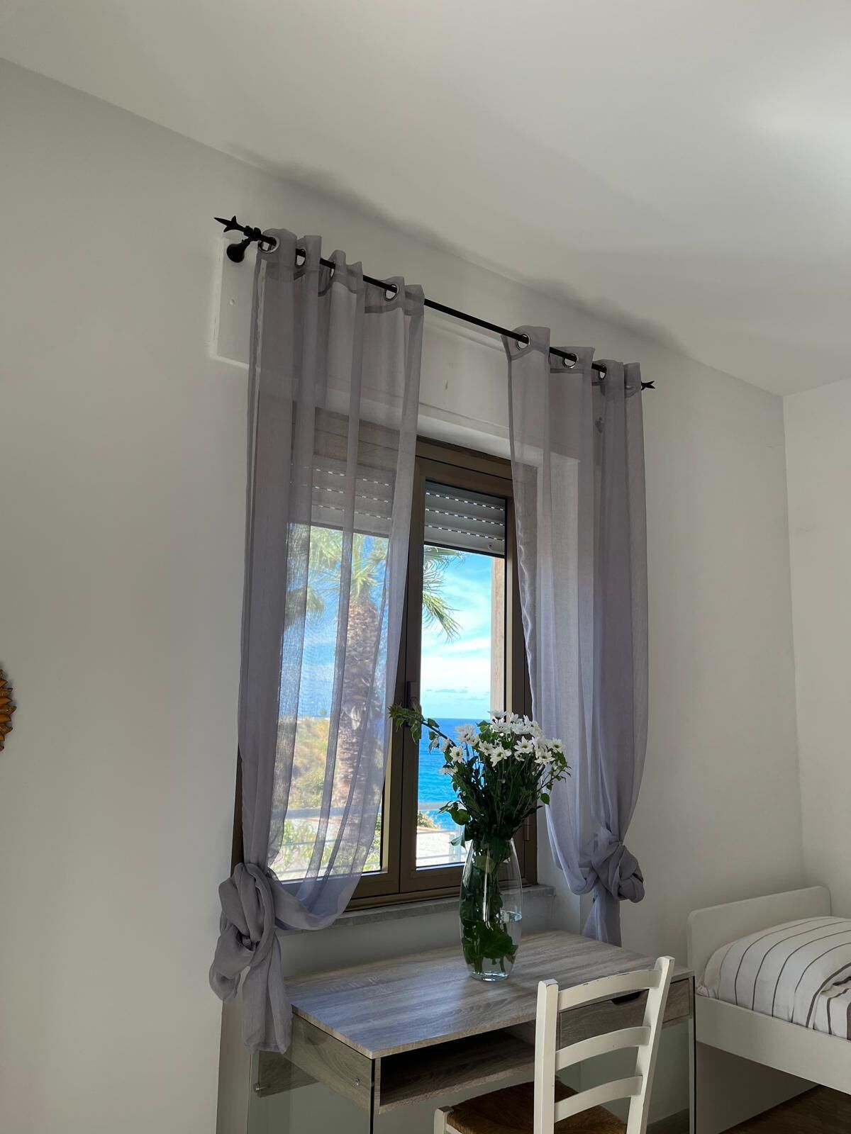 Un dormitorio con cama, mesita de noche, silla, ventana con cortinas y vista al mar.