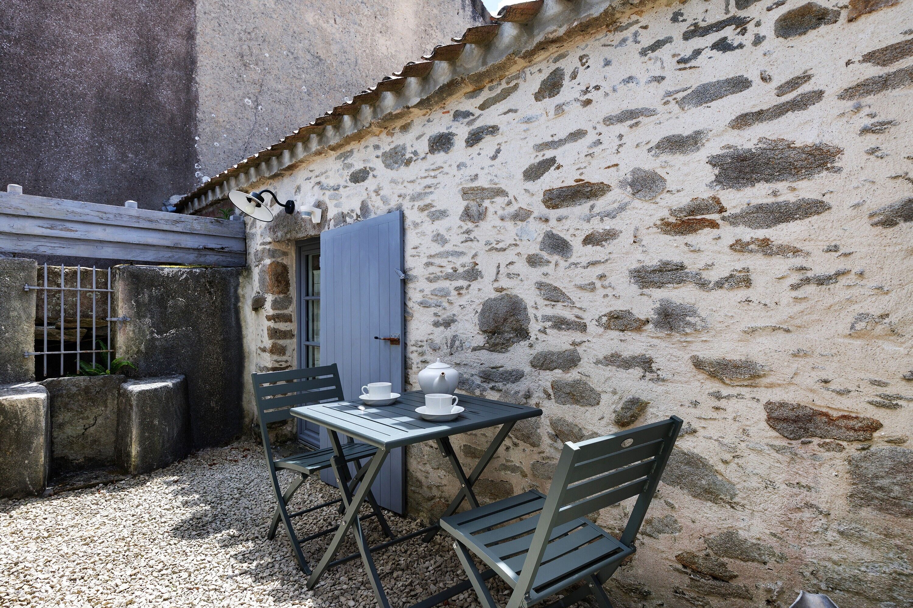 Un mur de pierre, une porte bleue, une table avec des chaises et un petit jardin.
