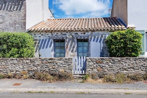 Un bâtiment aux murs de pierre, surmonté d’un toit en tuiles, avec deux fenêtres et une clôture blanche en bois.