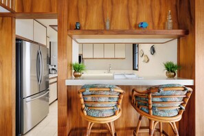 Une cuisine moderne avec un réfrigérateur en acier inoxydable, des armoires en bois, un plan de travail blanc et deux tabourets de bar dotés de coussins à motifs.