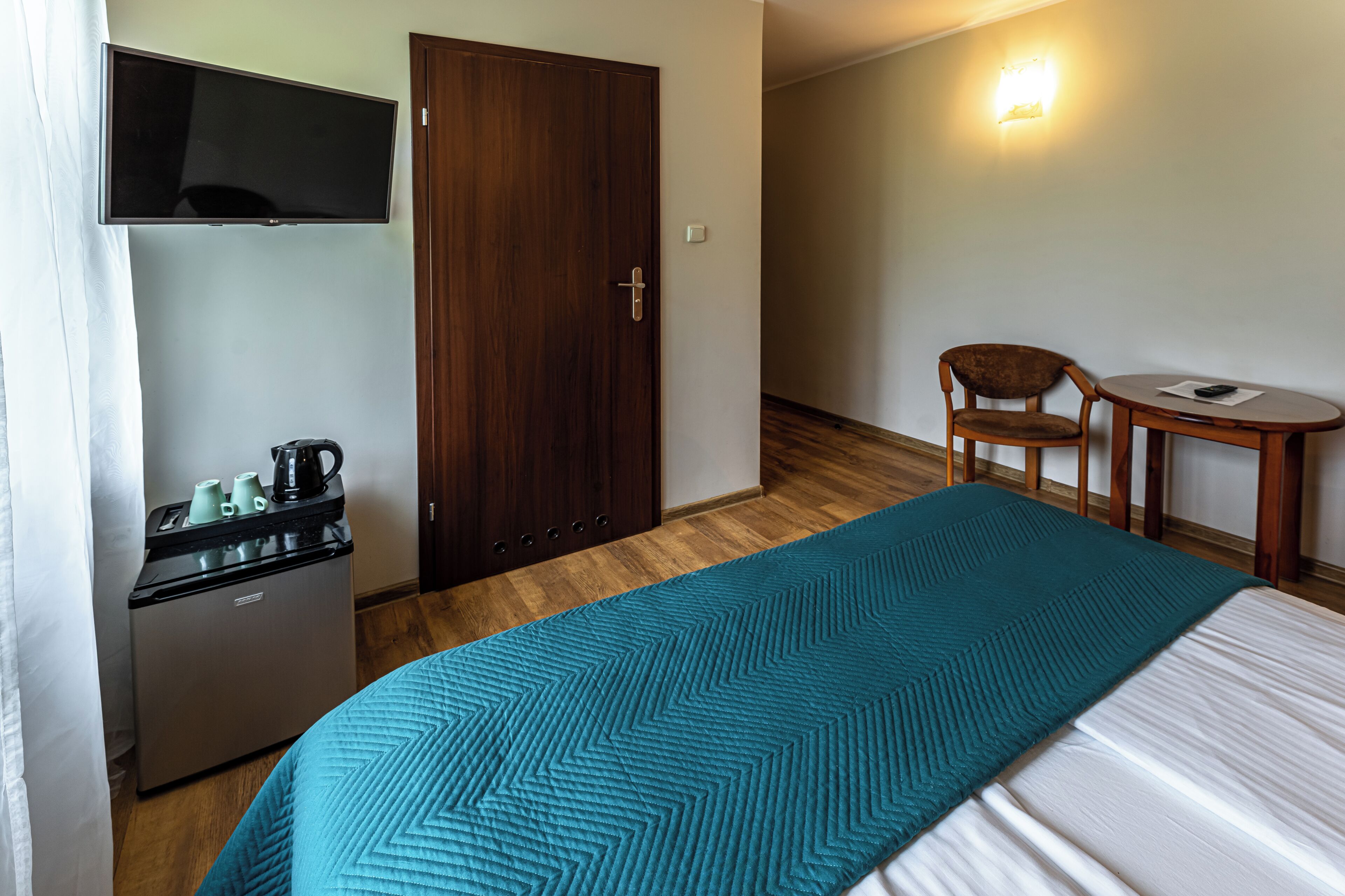 A hotel room with a bed, a desk with a chair, a TV mounted on the wall, and a small kitchenette.