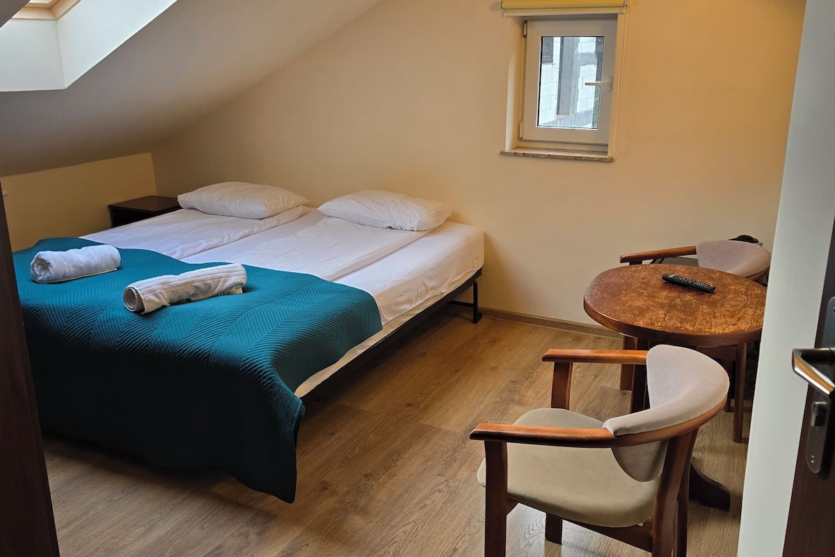 Une chambre avec un lit, une petite table et une chaise, un téléviseur fixé au mur et une fenêtre avec un store.