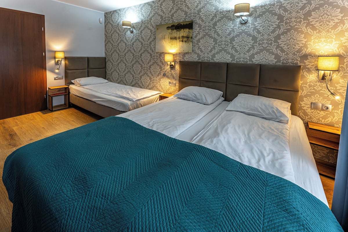 Une chambre d’hôtel avec deux lits, un grand lit recouvert d’une couverture turquoise, et deux tables de chevet avec des lampes.