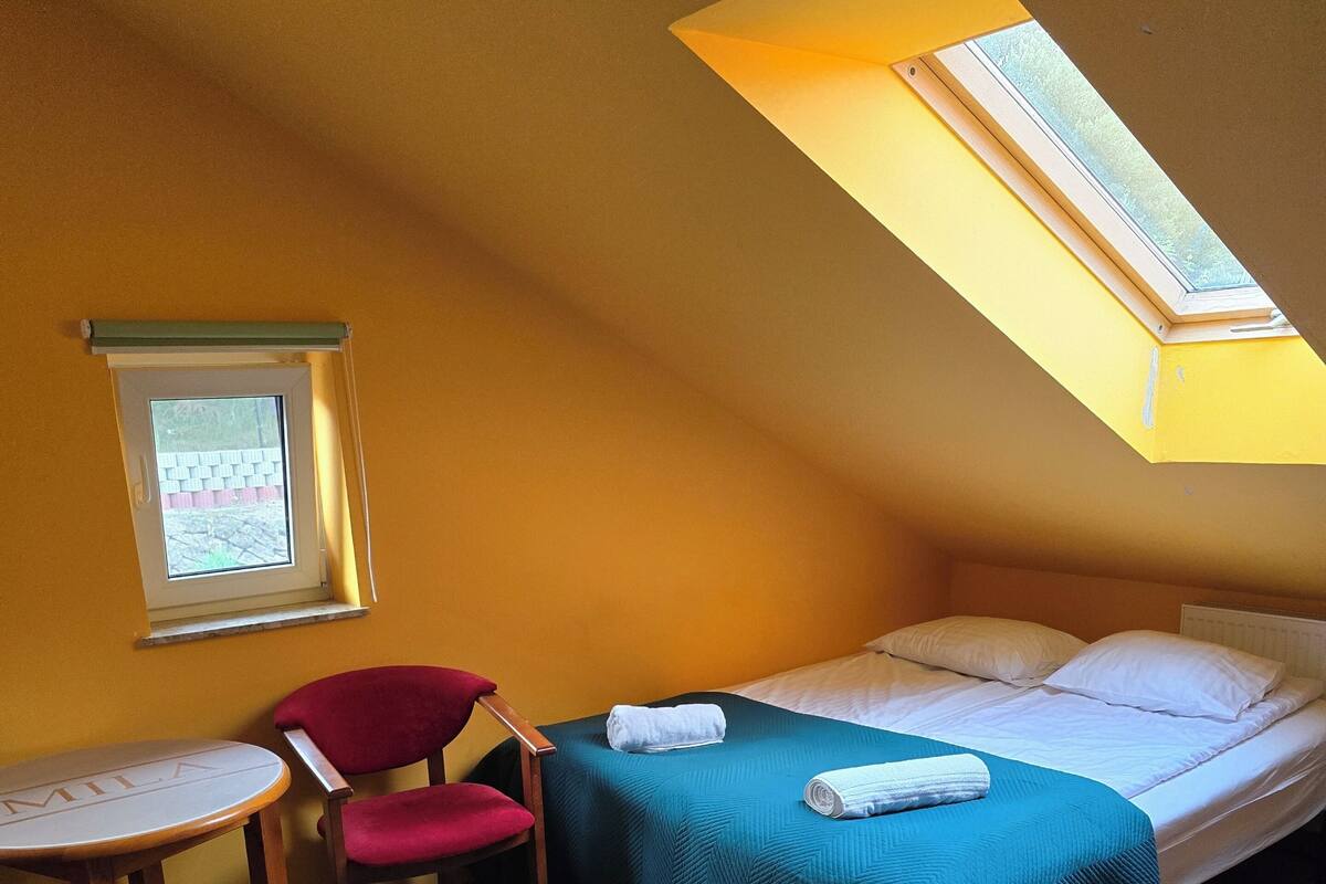 Un petit appartement d’une seule pièce, avec un plafond en pente, un lit, une chaise rouge, une petite table et une fenêtre.