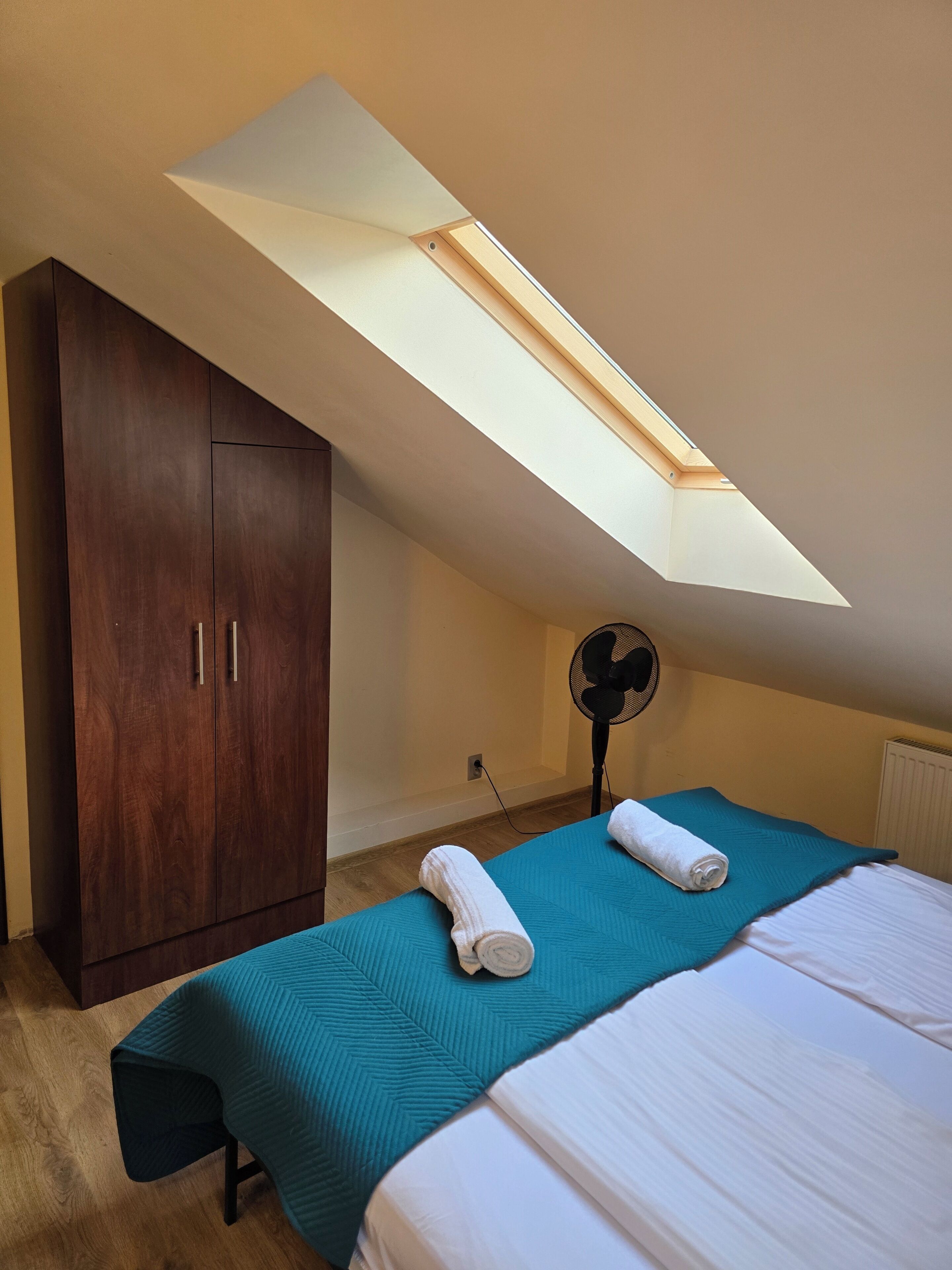 Ein Schlafzimmer mit Dachfenster, einem Bett mit petrolfarbener Bettdecke, einem Holzschrank und einem Standventilator.
