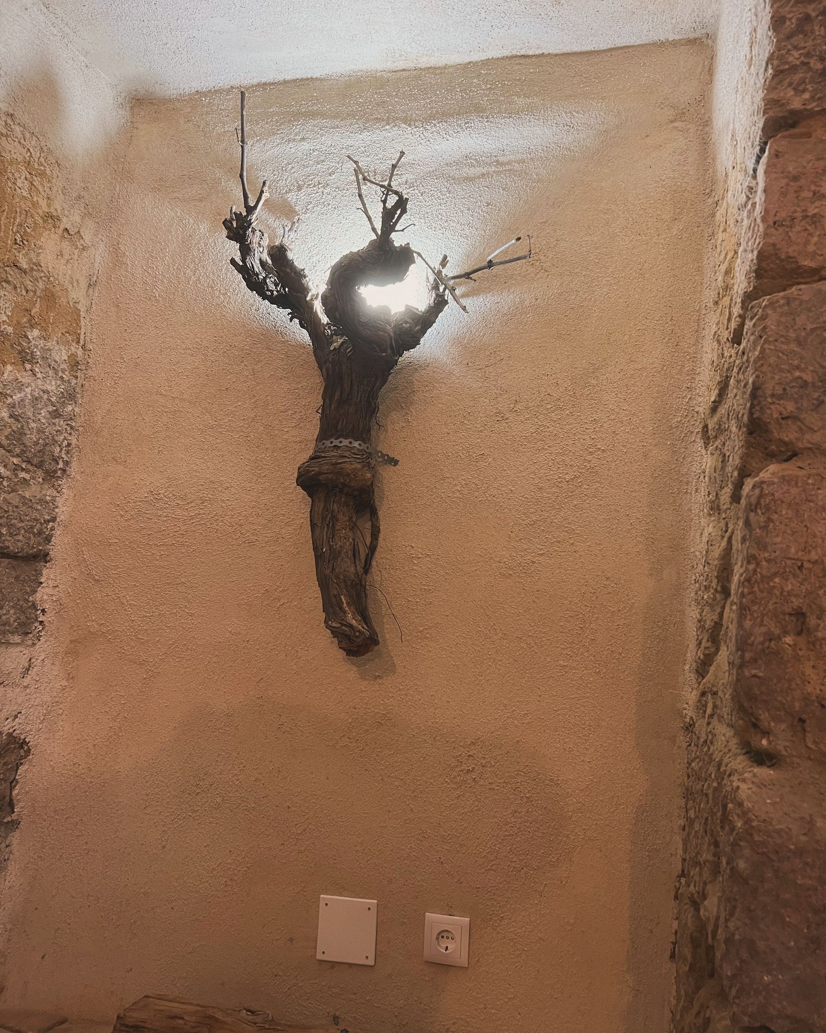 Eine an der Wand befestigte Skulptur mit baumartiger Struktur, möglicherweise aus Metall oder Stein, vor einer strukturierten, beige farbenen Wand.