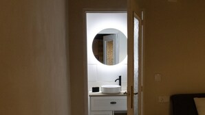 Passadís estret que condueix a un bany amb un mirall rodó, un lavabo i una tauleta de nit amb una planta.