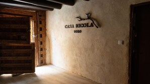 Un interior rústic amb una porta de fusta, un cartell fixat a la paret que diu "CASA NIGOLA TORO", i una estructura de bigues de fusta.