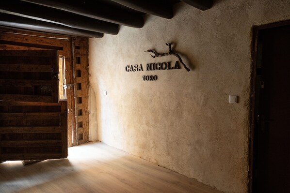 Un interior rústico con una puerta de madera, un letrero en la pared que dice "CASA NIGOLA TORO" y estructura de vigas de madera.