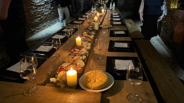 Una llarga taula de dinar arrematada amb espelmes, copes de vi i plats de menjar, que suggereix una experiència gastronòmica formal en una estança poc il·luminada amb parets de maó.