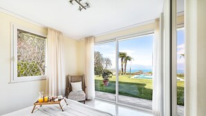 Un dormitorio con cama, mesita de noche con productos para el desayuno, una silla y un amplio ventanal corredero que da a un jardín con piscina.