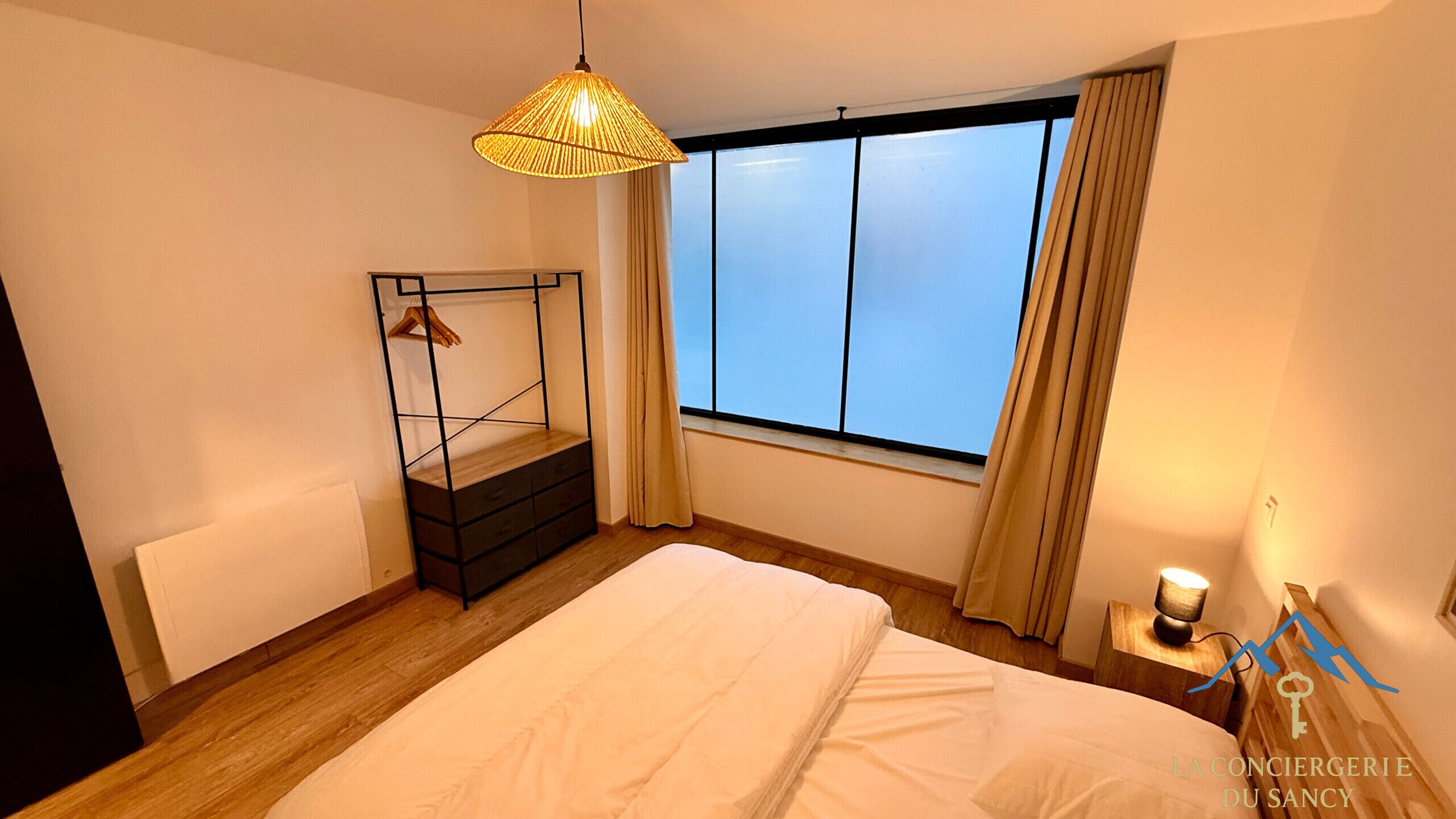 Un dormitorio con cama, mesita de noche, lámpara colgante, ventana con cortinas y estante en la pared.