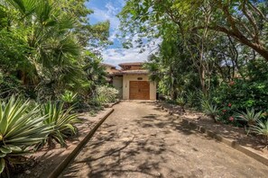 A pathway leading to a building with a tiled roof, surrounded by various plants and trees. - Naka-Mar Beachfront Villa (Las Golondrinas , Hacienda Pinilla)
