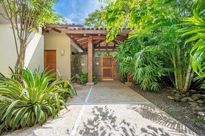 A covered outdoor area with a stone wall, a wooden door, and various tropical plants. - Naka-Mar Beachfront Villa (Las Golondrinas , Hacienda Pinilla)