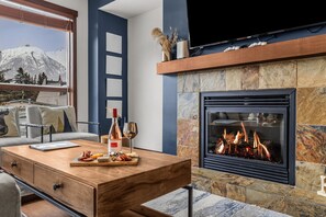 A modern living room with a fireplace, a sofa, a coffee table with snacks, and a view of mountains through a large window. - Downtown |Sleeps 10 | Walk to shopping | Restaurants| Creekside trails and Ponds (Canmore)
