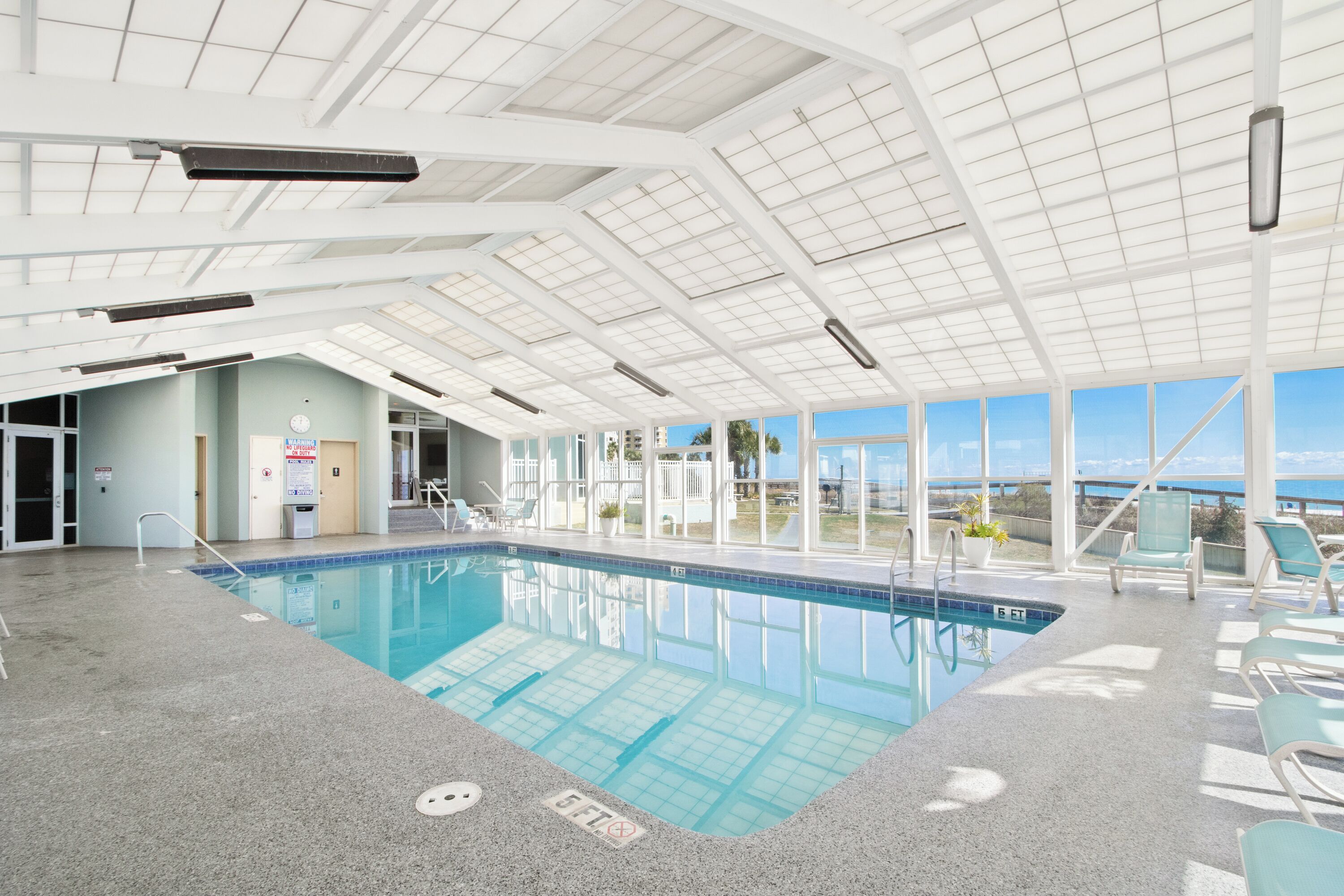An indoor swimming pool with clear blue water, surrounded by lounge chairs and a view of the sea through large windows.
