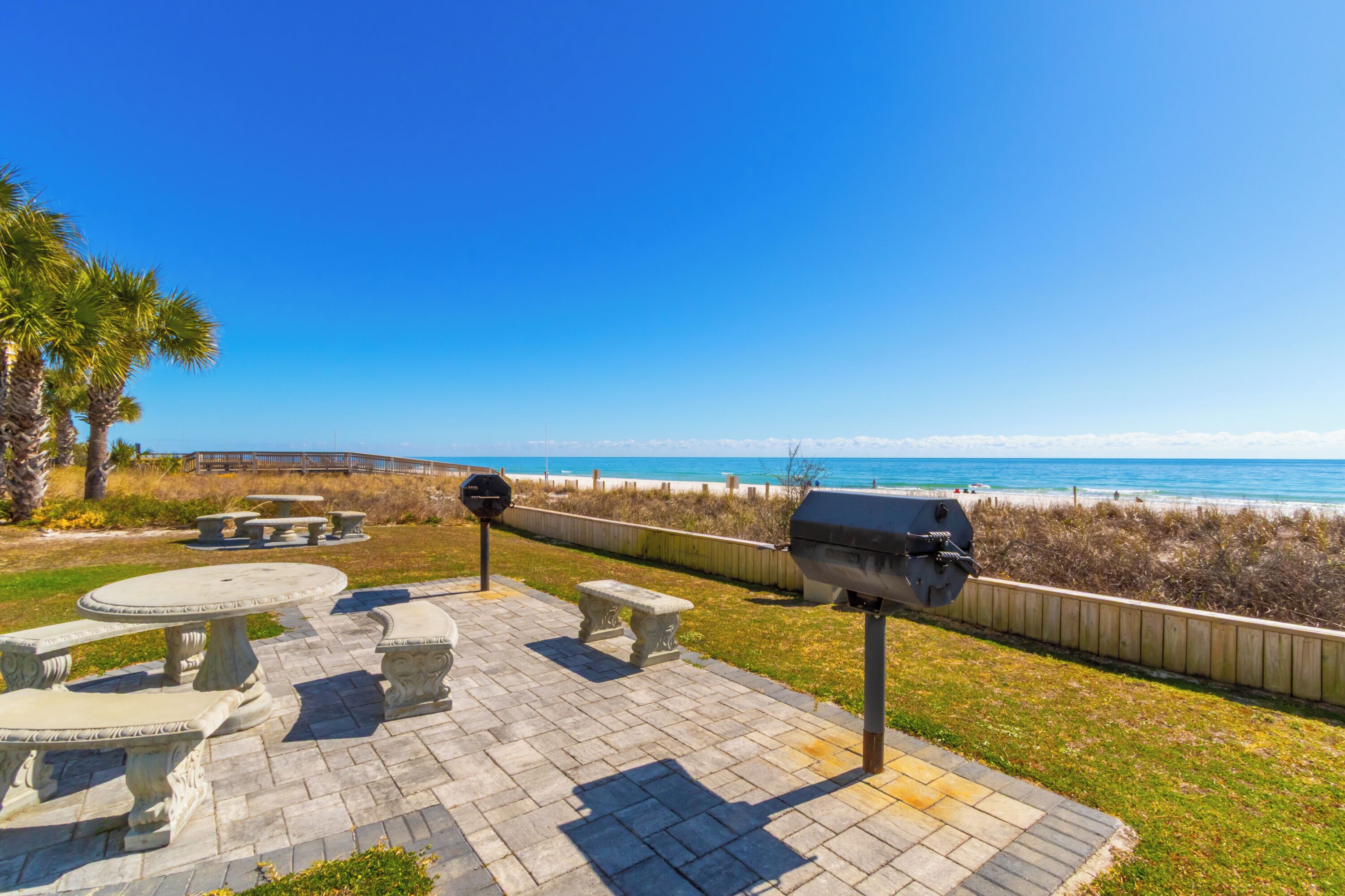 A patio with stone tables and benches, a barbecue grill, and a view of the beach and ocean.